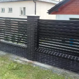 Nowoczesne ogrodzenie panelowe z czarnych, poziomych listew, osadzone na słupkach z czarnej cegły klinkierowej, z widoczną skrzynką gazową po lewej stronie.