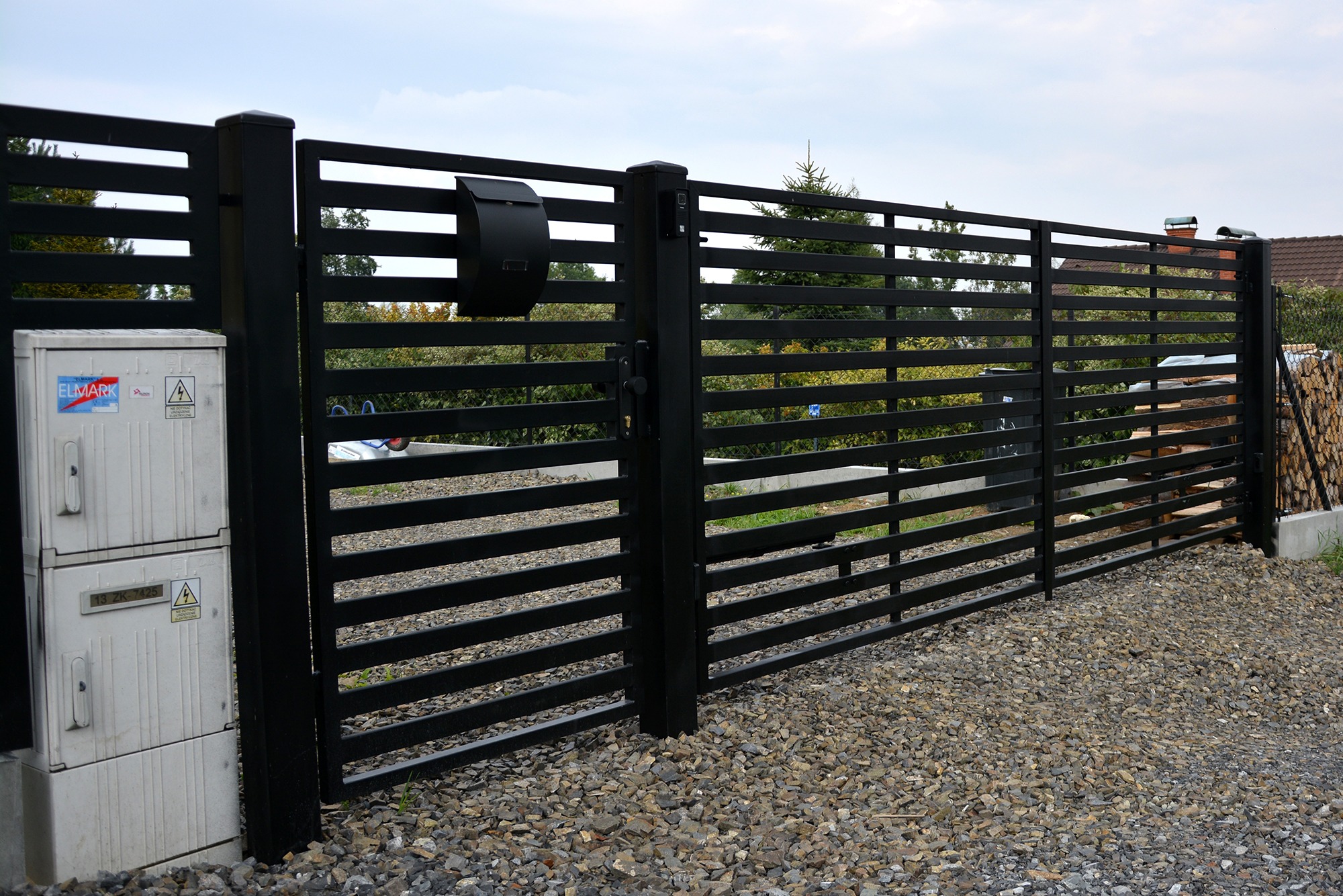 Nowoczesna, pozioma brama wjazdowa z czarnych, metalowych profili w Andrychowie, z zamontowaną skrzynką na listy, widoczna skrzynka elektryczna i ułożone drewno na posesji.