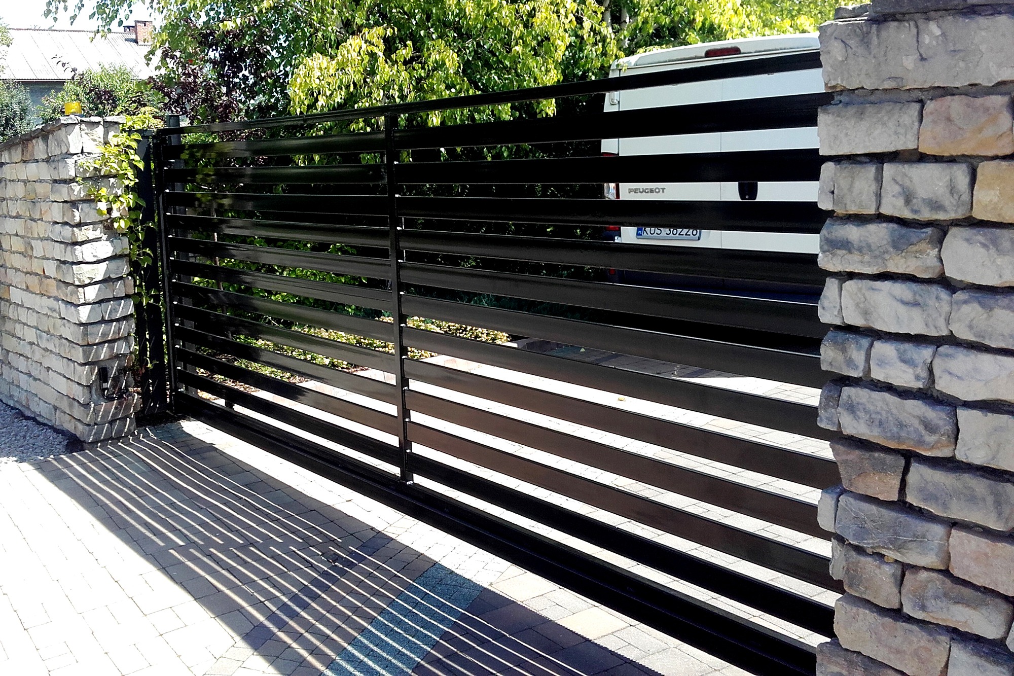 Nowoczesna, czarna brama wjazdowa z poziomych profili, zamontowana między kamiennymi słupkami, z widocznym fragmentem zaparkowanego białego samochodu w tle.