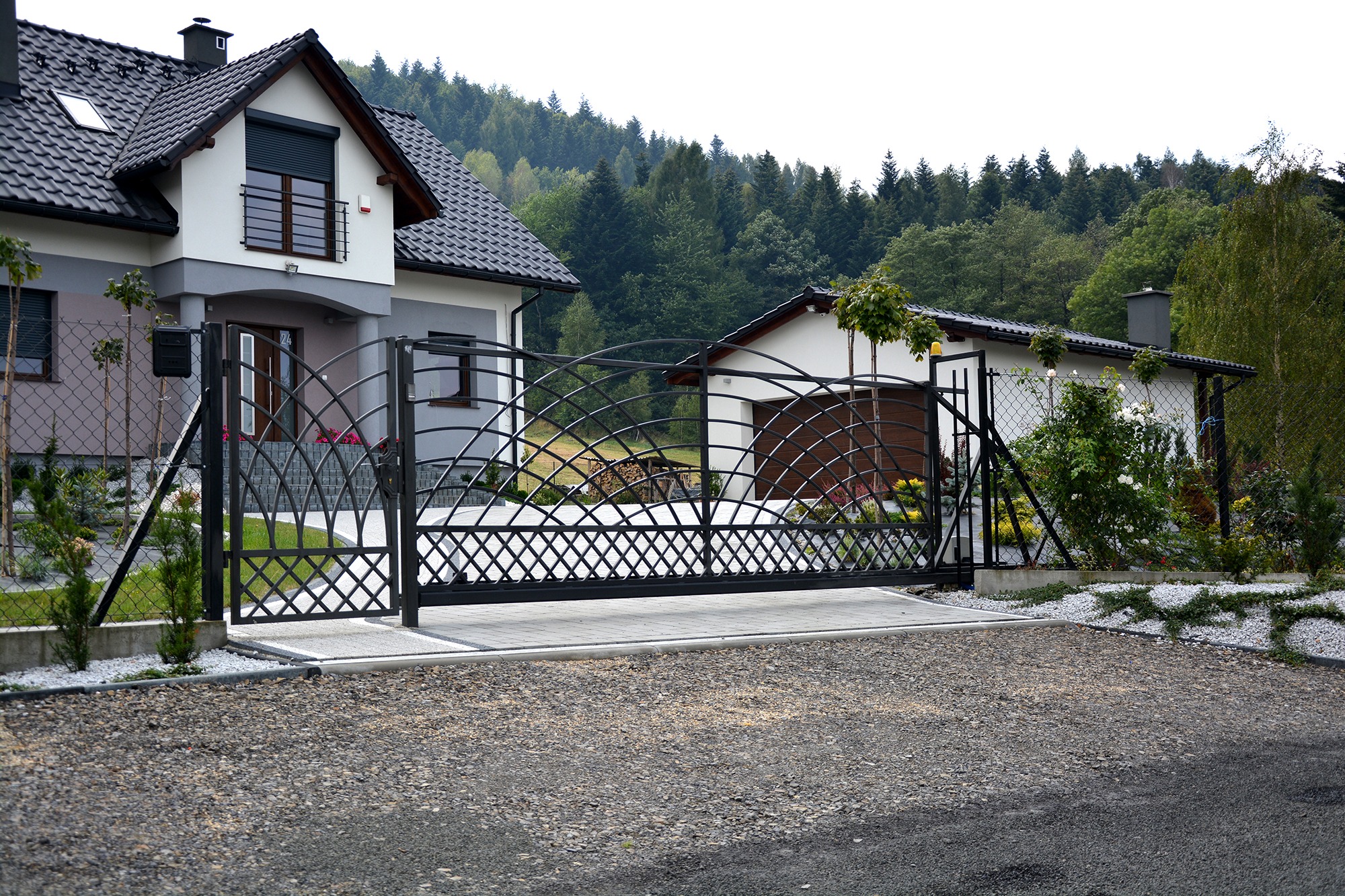 Czarna, metalowa brama wjazdowa z dekoracyjnym wzorem na tle domu jednorodzinnego z garażem i zielenią.