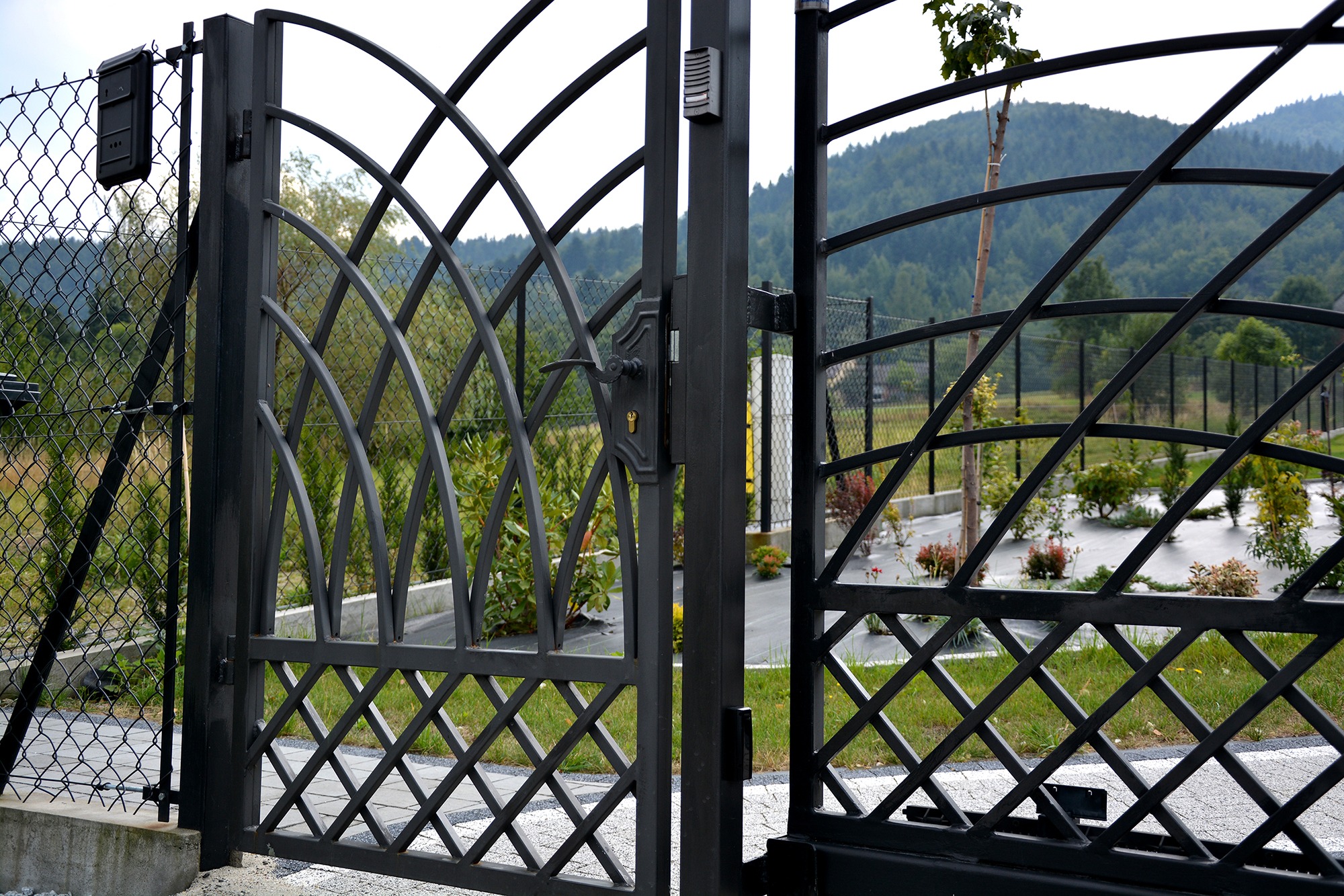 Otwarta, czarna, metalowa brama wjazdowa z ozdobnym wzorem kraty i łuków, z domofonem i klamką, widok na ogród z elementami małej architektury i zielone wzgórza w tle.