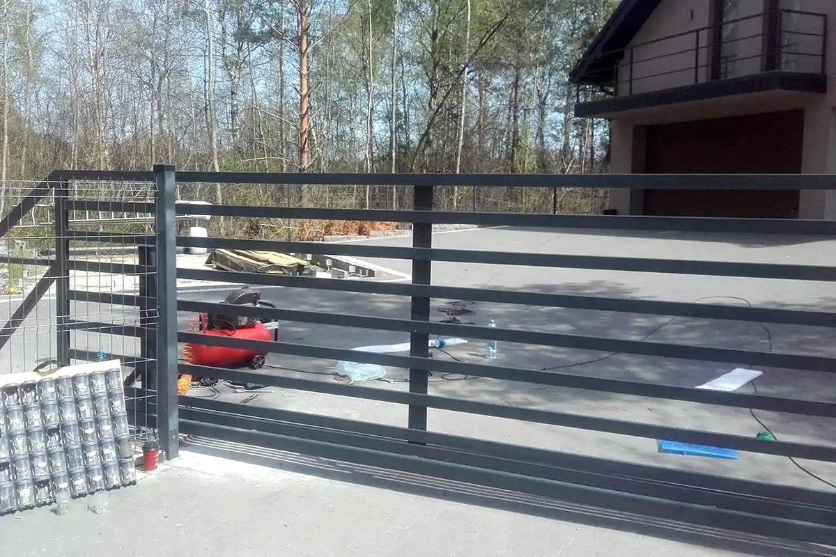 Metalowa brama wjazdowa przesuwna w trakcie montażu, widoczny kompresor, puszki farby i narzędzia na betonowym podłożu, w tle dom jednorodzinny i drzewa.