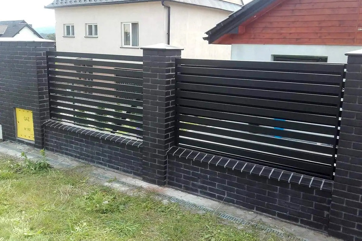 Nowoczesne ogrodzenie panelowe z czarnych, poziomych listew, osadzone na słupkach z czarnej cegły klinkierowej, z widoczną skrzynką gazową po lewej stronie.