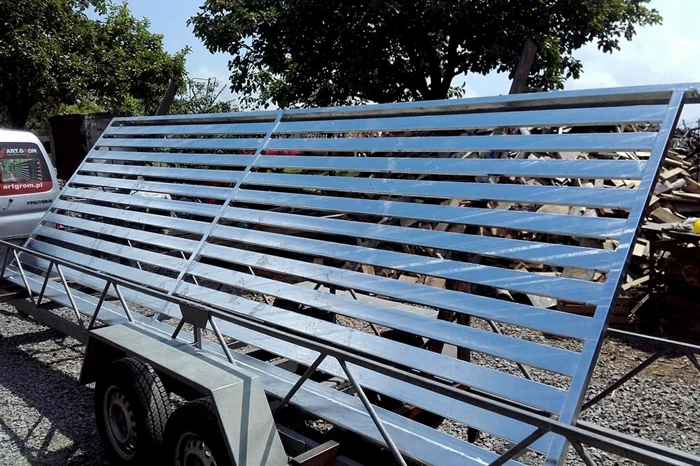 Nowa, ocynkowana brama wjazdowa z poziomymi szczeblami, przewożona na przyczepie samochodowej, z widocznym stosem drewna w tle.