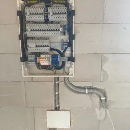 Rozdzielnica elektryczna z zabezpieczeniami zamontowana na surowej ścianie z bloczków betonowych, widoczne przewody w peszlu i listwy zaciskowe.