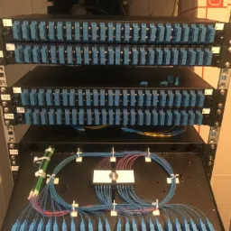 Panel krosowniczy z licznymi złączami światłowodowymi, uporządkowane kable w kolorach niebieskim i czerwonym, zamocowane uchwytami, widoczne oznaczenia portów.