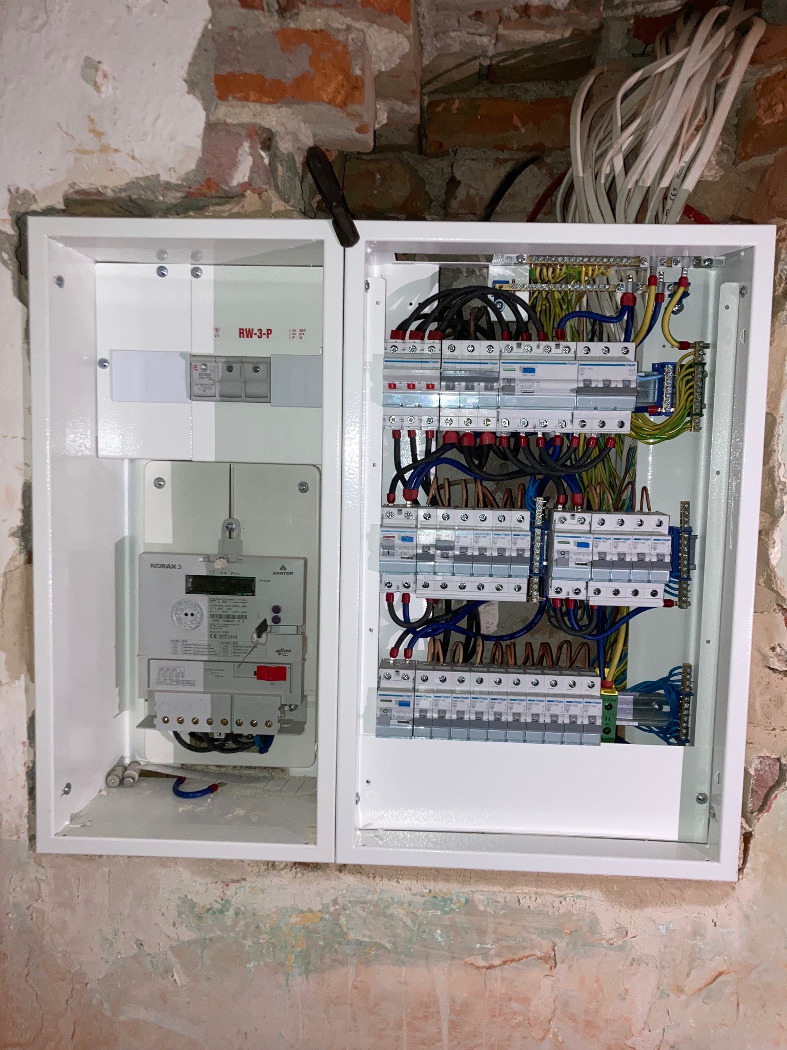 Rozdzielnica elektryczna z licznikiem i zabezpieczeniami, zamontowana na surowej ścianie z cegły i resztkami farby, widoczne kable i przewody.