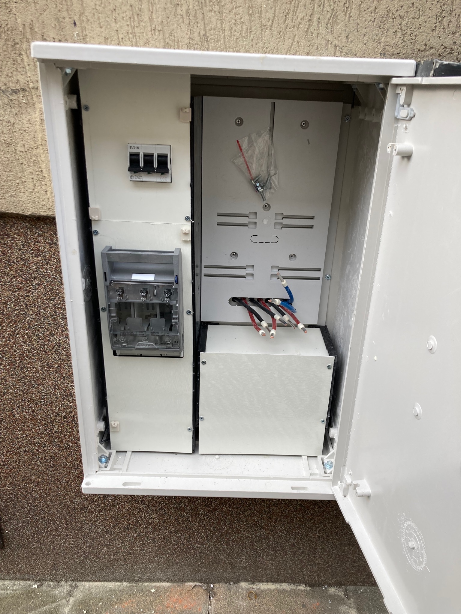 Otwarta, biała skrzynka elektryczna na elewacji budynku, widoczne zabezpieczenia, kable i puste miejsce na licznik.