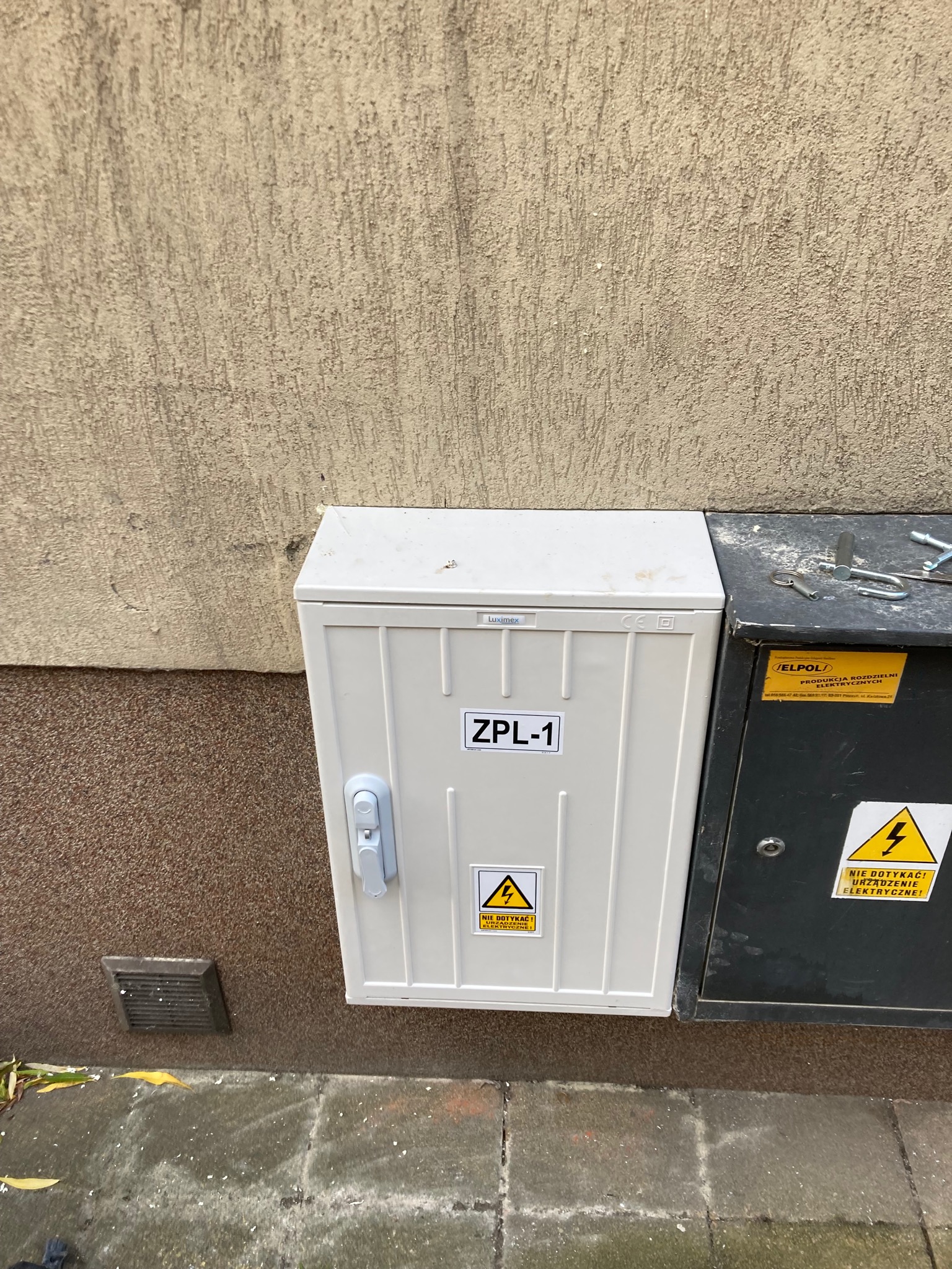 Biała skrzynka elektryczna ZPL-1 zamontowana na ścianie budynku, obok ciemniejszej skrzynki z ostrzeżeniem o napięciu. Widoczne narzędzia na górze ciemnej skrzynki.
