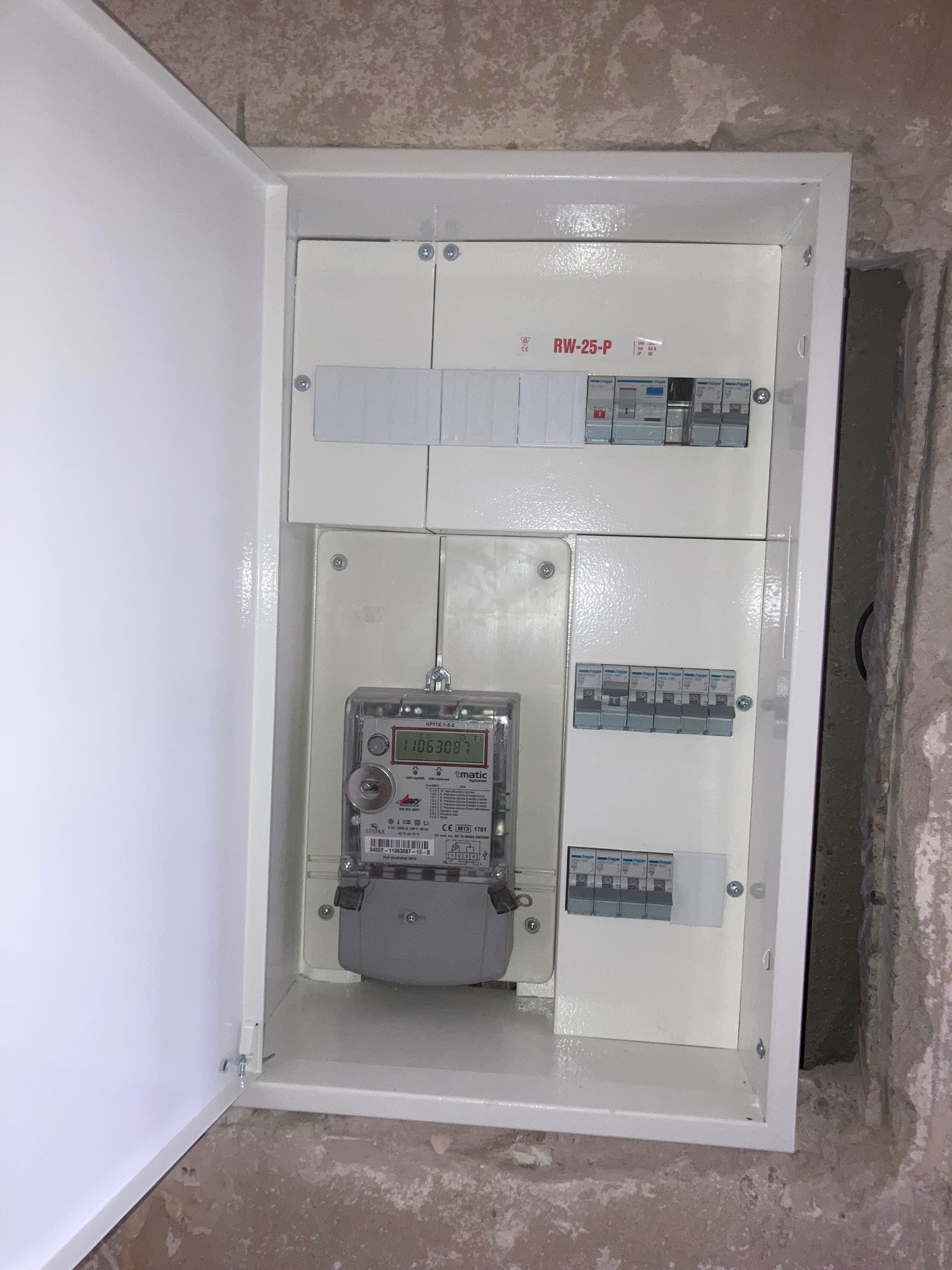 Otwarta skrzynka elektryczna z licznikiem i zabezpieczeniami na tle surowej ściany w trakcie remontu.