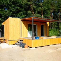 Nowoczesny, parterowy budynek z jasnego drewna z płaskim dachem i zadaszonym tarasem, częściowo wykończony, z widocznymi schodami wejściowymi, stojący na piaszczystym terenie z lasem w tle.