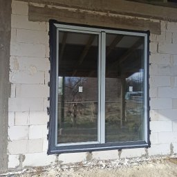 KAMBUD Kamil Laskowski - Dwukomorowe okno aluminiowe z szarą uszczelką zamontowane w surowym murze z betonu komórkowego; widoczne drewniane elementy konstrukcji dachu wewnątrz budynku.