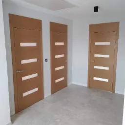 Trzy zamontowane drzwi wewnętrzne z poziomymi przeszkleniami w jasnym pomieszczeniu z szarą podłogą i białymi ścianami, widoczne czarne lampy sufitowe.