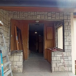 KAMBUD Kamil Laskowski - Wejście do domu w trakcie remontu: widoczne ceglane wykończenie portalu, częściowo zamontowane okno, drzwi opierające się o ścianę, drabina i materiały budowlane. Wnętrze domu z boazerią w tle.