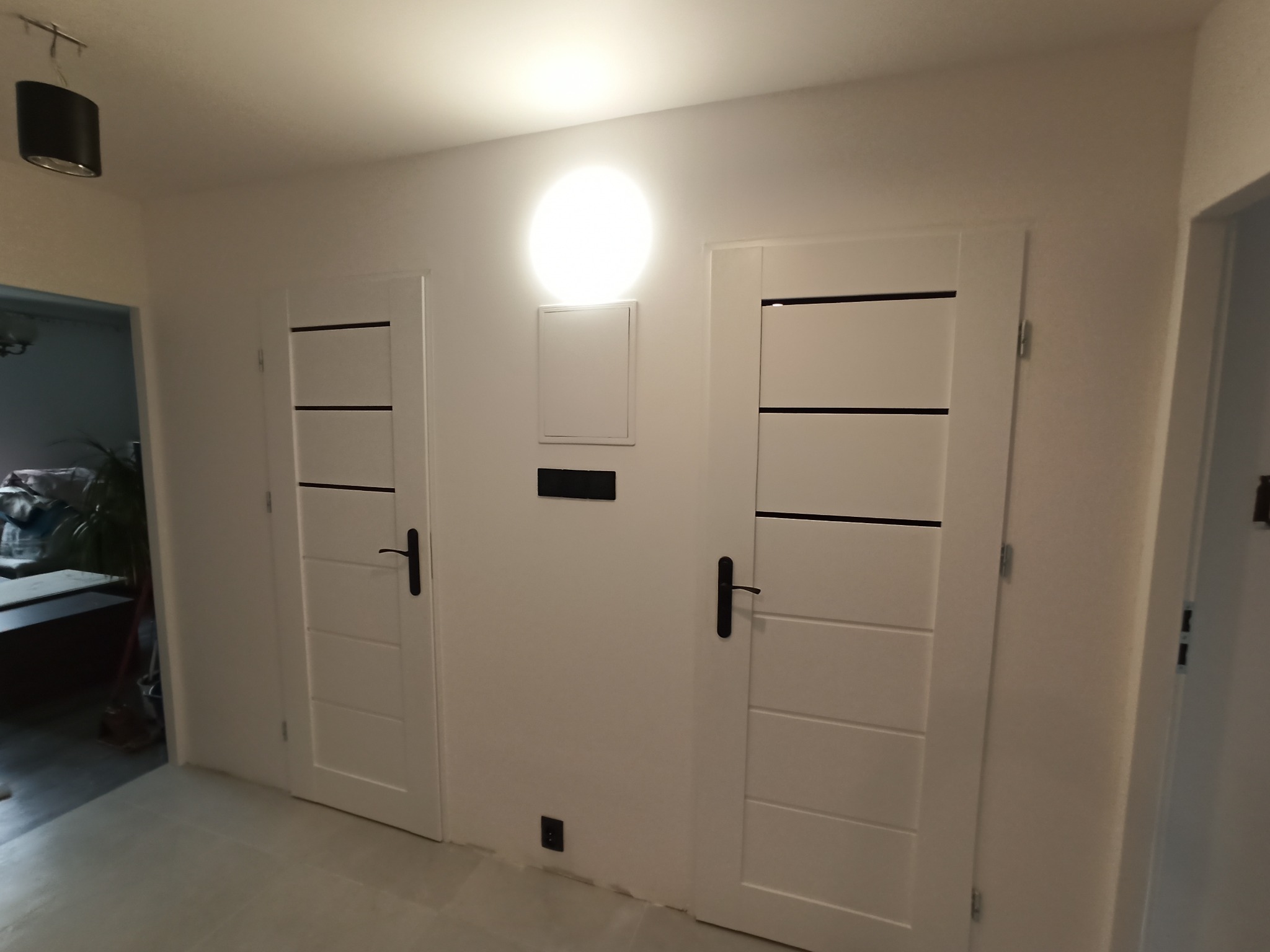 Dwa białe, nowoczesne drzwi wewnętrzne z poziomymi, czarnymi wstawkami i czarnymi klamkami, zamontowane w jasnym korytarzu z szarą podłogą, oświetlone centralnym okrągłym plafonem.