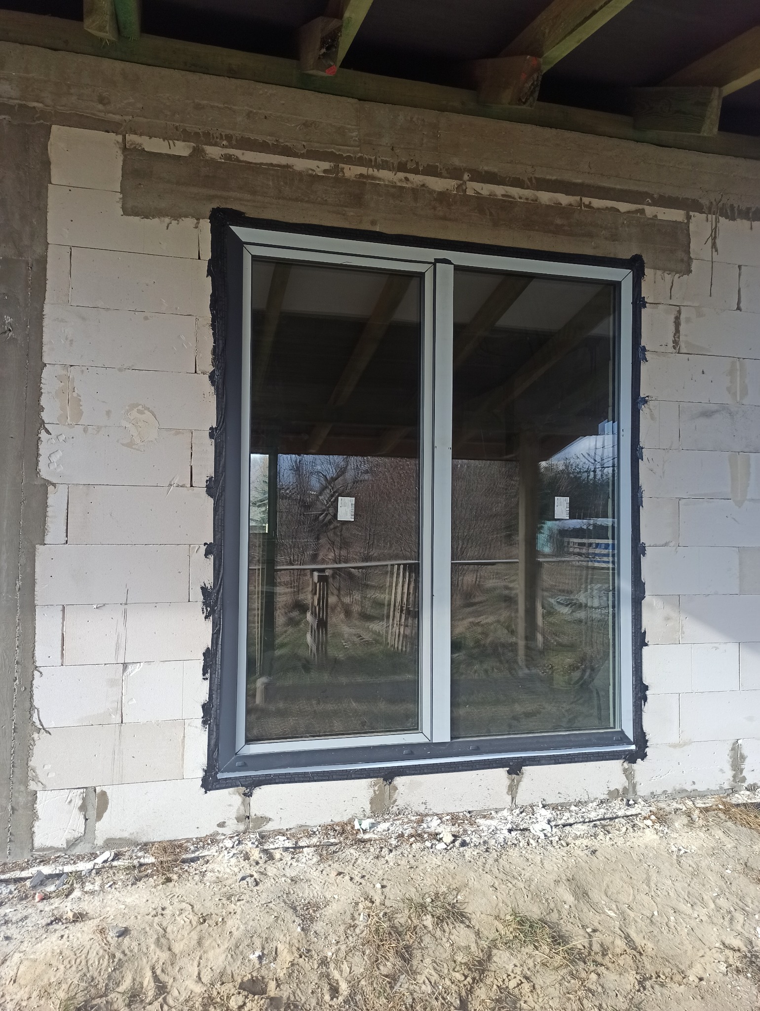 Dwukomorowe okno aluminiowe z szarą uszczelką zamontowane w surowym murze z betonu komórkowego; widoczne drewniane elementy konstrukcji dachu wewnątrz budynku.