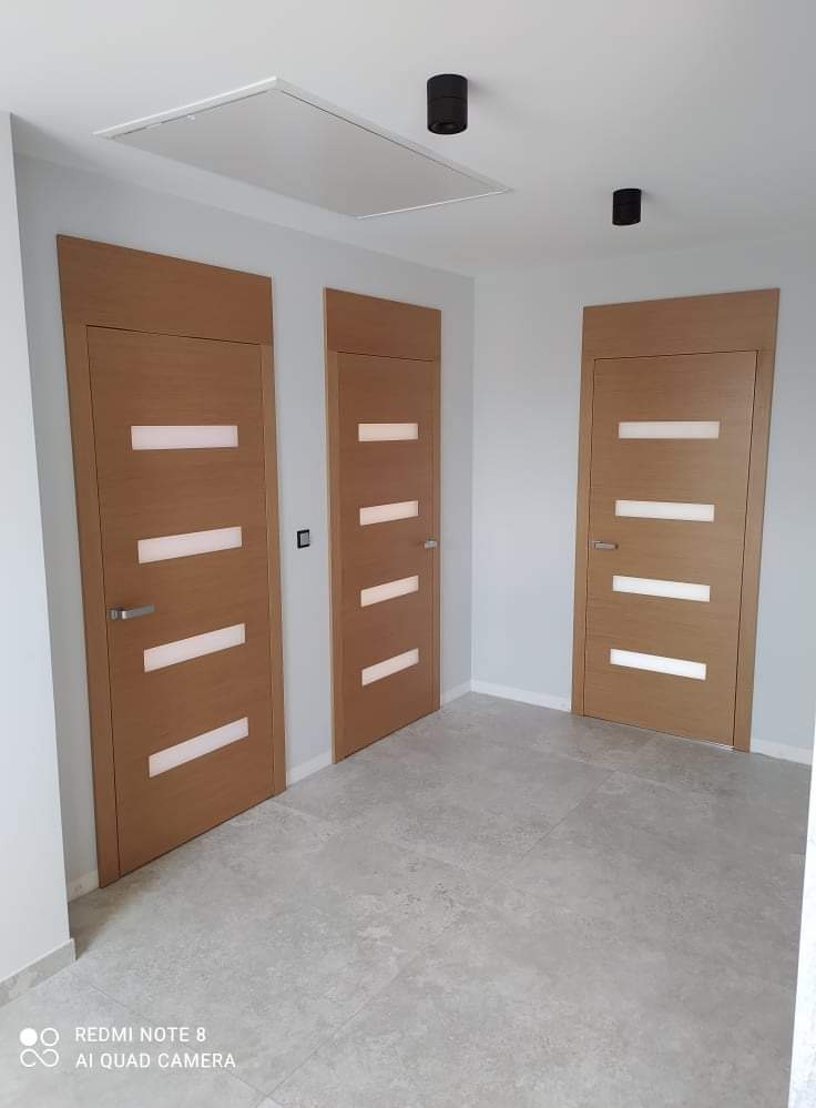 Trzy zamontowane drzwi wewnętrzne z poziomymi przeszkleniami w jasnym pomieszczeniu z szarą podłogą i białymi ścianami, widoczne czarne lampy sufitowe.