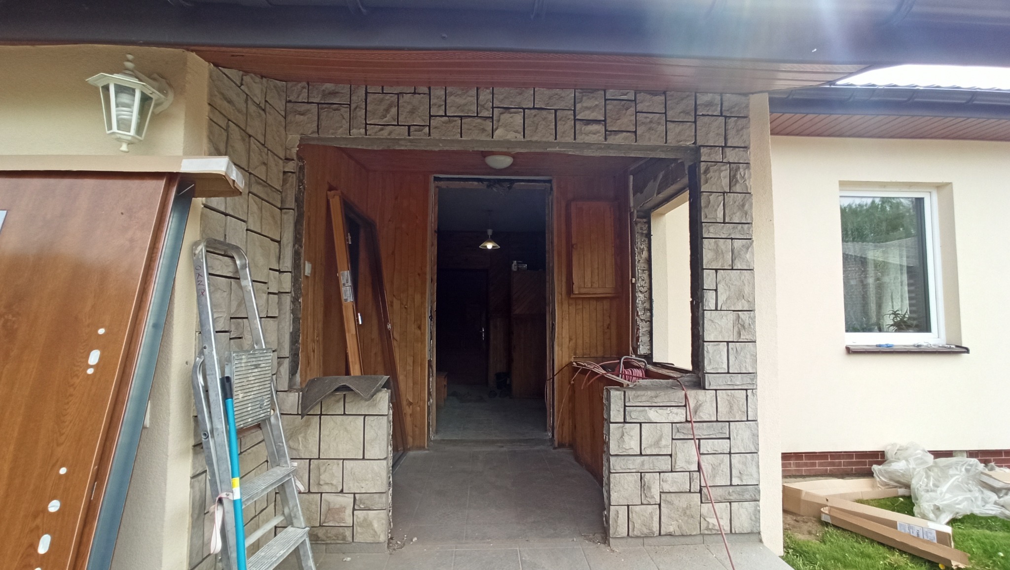 Wejście do domu w trakcie remontu: widoczne ceglane wykończenie portalu, częściowo zamontowane okno, drzwi opierające się o ścianę, drabina i materiały budowlane. Wnętrze domu z boazerią w tle.