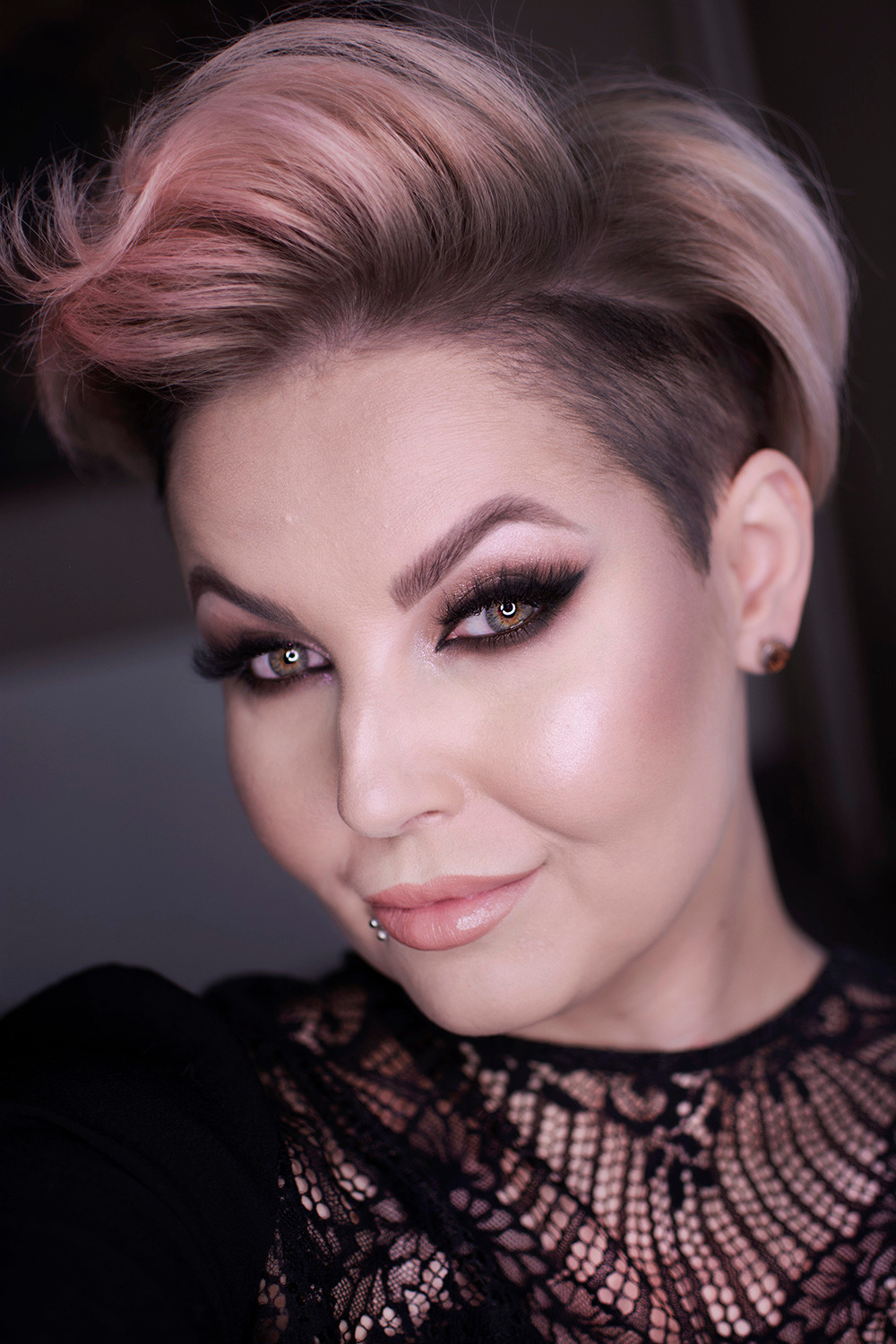 Portret kobiety z profesjonalnym makijażem smokey eye, podkreślonymi brwiami, błyszczącymi ustami i asymetryczną fryzurą w odcieniach blondu i różu, noszącej czarną koronkową bluzkę i kolczyk...