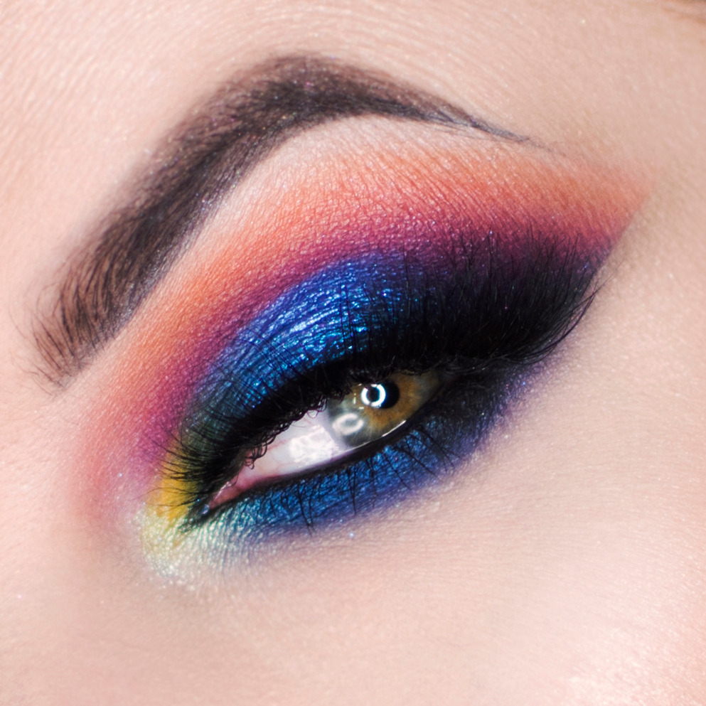 Intensywny makijaż oka z użyciem cieni w odcieniach pomarańczy, różu, fioletu i błyszczącego granatu, podkreślony czarnym eyelinerem i sztucznymi rzęsami, widoczne delikatne załamania na powiece.