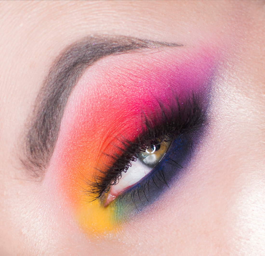 Zbliżenie oka z intensywnym, wielobarwnym cieniowaniem: żółty, pomarańczowy, czerwony, różowy, fioletowy i granatowy. Wyraźnie podkreślone rzęsy i brwi. Skóra o delikatnej fakturze.