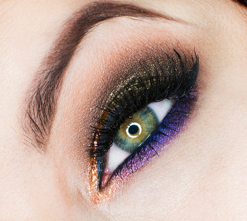 Zbliżenie oka z intensywnym, artystycznym zdobieniem: złoty brokat na powiece górnej, fioletowy cień na dolnej powiece, wydłużone, czarne rzęsy i wyraźnie zaznaczone brwi.
