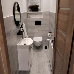 Nowoczesna, wąska łazienka w szaro-białej kolorystyce z wiszącą toaletą, umywalką z szafką, grzejnikiem drabinkowym i okrągłym lustrem z czarną ramą, widok zza drewnianych drzwi.