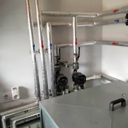 Instalacja grzewcza z izolowanymi rurami, pompami obiegowymi i sterownikiem, zamontowana na białej ścianie w pomieszczeniu technicznym.