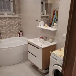 Jasna łazienka z narożną wanną, umywalką z szafką i pralką Samsung, widoczne kosmetyki na półce i ręczniki w lustrze.