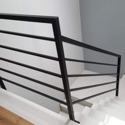 Zakład Tokarsko Ślusarski Igański Mirosław - Minimalistyczna, czarna balustrada z poziomych prętów zabezpieczająca białe schody w nowoczesnym wnętrzu z szarą ścianą.