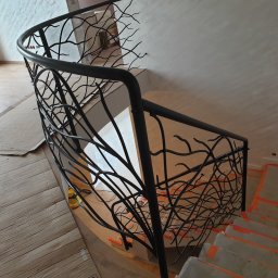 Zakład Tokarsko Ślusarski Igański Mirosław - Czarna, metalowa balustrada o wzorze przypominającym gałęzie drzewa, zamontowana na schodach w trakcie prac wykończeniowych, zabezpieczonych pomarańczową taśmą malarską.
