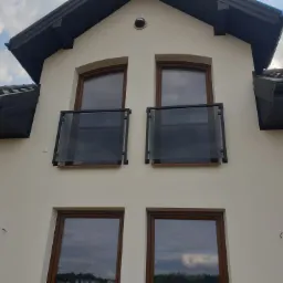 Elewacja domu z dwoma balkonami z balustradami szklanymi na piętrze i dwoma oknami na parterze, widok z dołu na tle błękitnego nieba z chmurami.