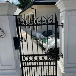 Czarna, metalowa brama wjazdowa z dekoracyjnymi elementami, osadzona w białych, murowanych słupkach, prowadząca na posesję z beżowym domem i brukowanym podjazdem.