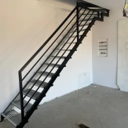 Metalowe schody z kratowymi stopniami i czarną balustradą, zamontowane w surowym wnętrzu z widoczną instalacją elektryczną i śladami po pracach wykończeniowych.