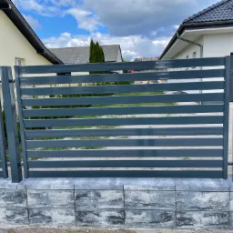 Nowoczesne, poziome ogrodzenie metalowe w kolorze grafitowym, osadzone na betonowej podmurówce, z widocznymi domami w tle.