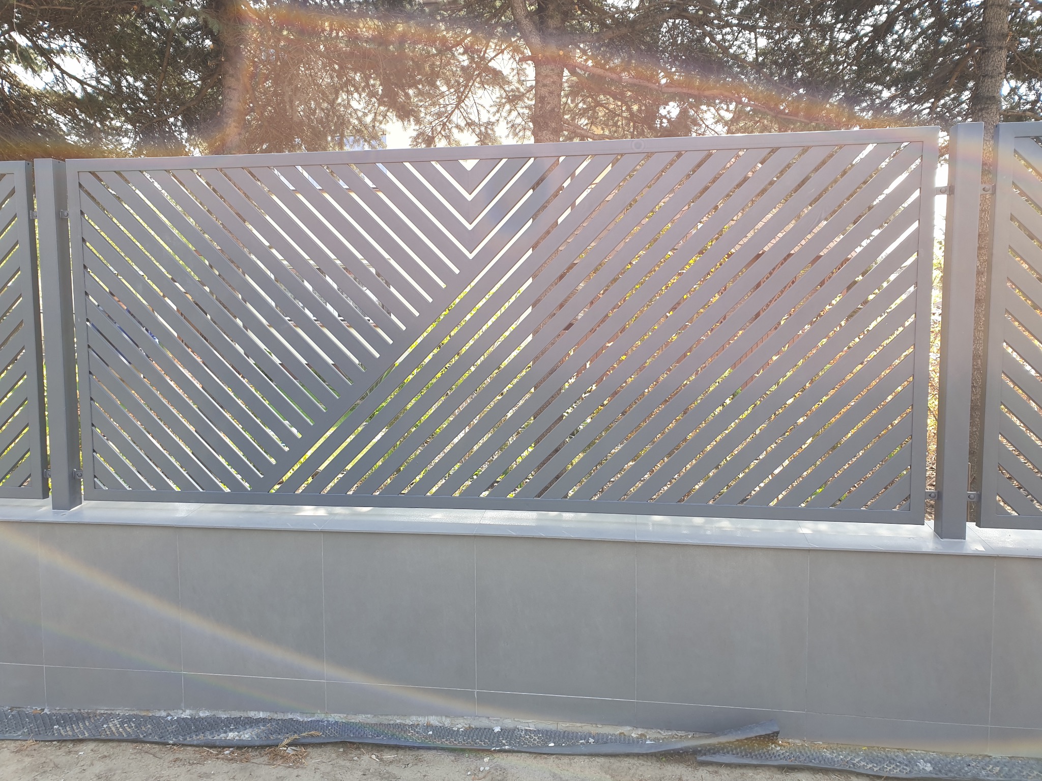 Nowoczesne, szare ogrodzenie z ukośnymi, ażurowymi wzorami, zamontowane na niskim, płytkowanym murku, z widocznym drzewem w tle i refleksami słonecznymi.