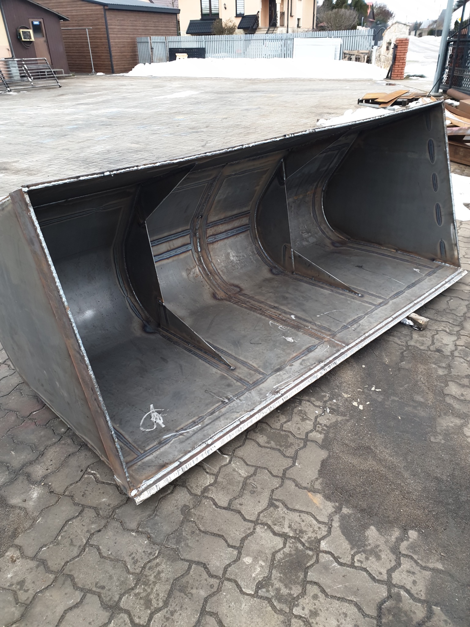 Surowy, spawany metalowy kosz ładowarki na zewnątrz, widoczne spawy i konstrukcja wzmacniająca, na tle kostka brukowa i zabudowania.