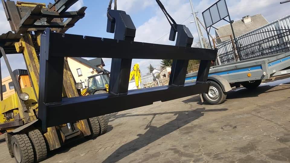 Czarny, metalowy element konstrukcyjny bramy transportowany przez wózek widłowy, w tle widoczna przyczepa z ogrodzeniem i maszyny budowlane.