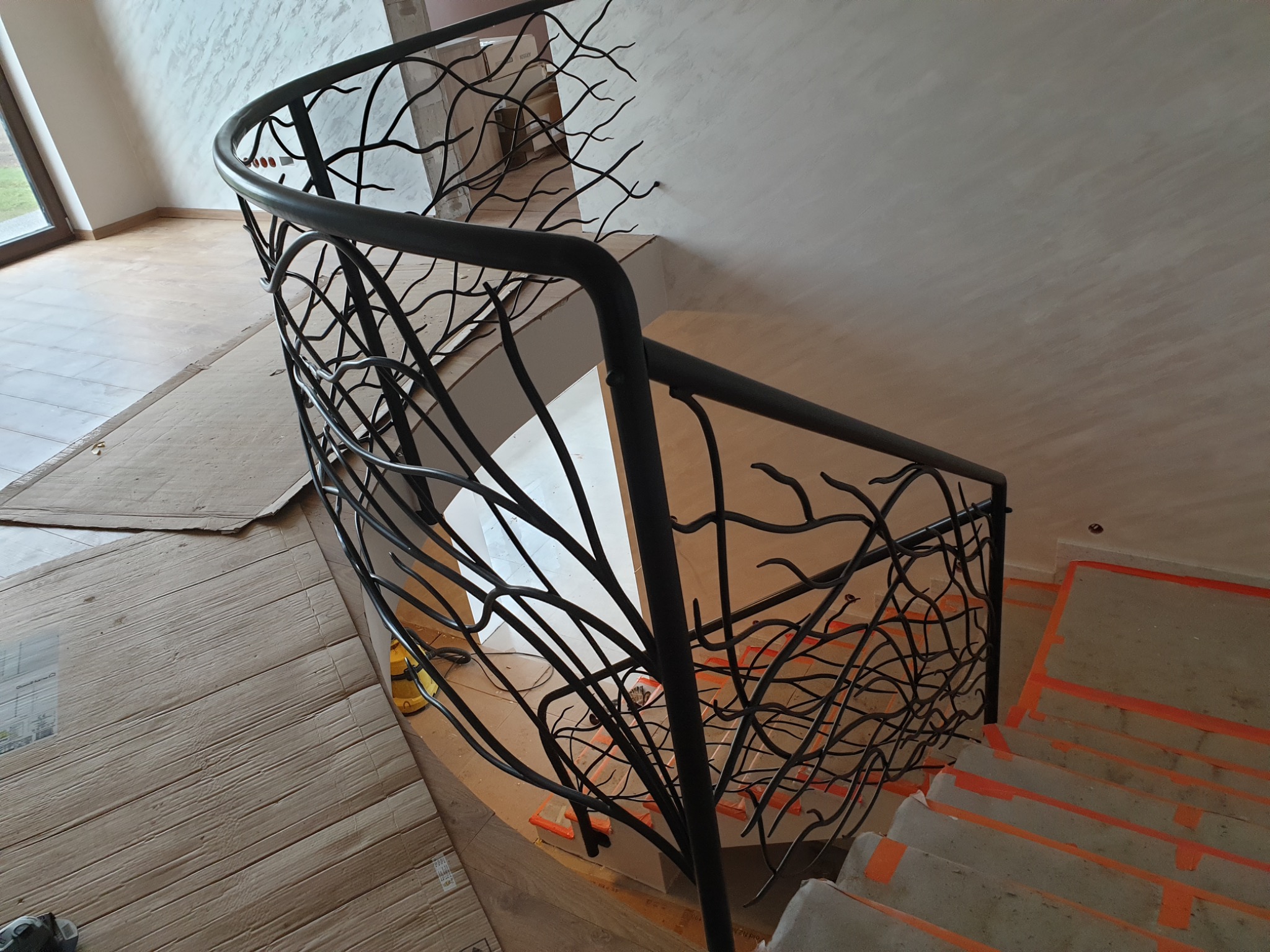 Czarna, metalowa balustrada o wzorze przypominającym gałęzie drzewa, zamontowana na schodach w trakcie prac wykończeniowych, zabezpieczonych pomarańczową taśmą malarską.