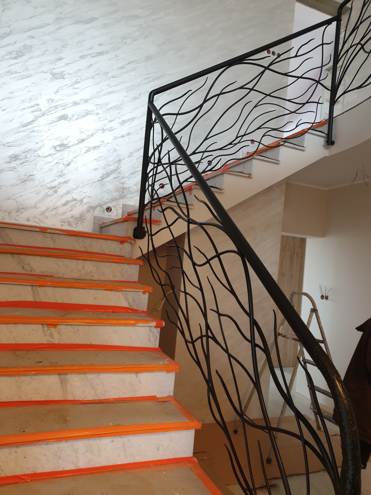 Marmurowe schody z czarną, dekoracyjną balustradą o motywie gałęzi, zabezpieczone folią ochronną podczas remontu.