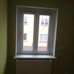 Mal-Bud - Białe, dwuskrzydłowe okno PCV z widokiem na blok mieszkalny, zamontowane w pokoju o ścianach pomalowanych na oliwkowy kolor, z grzejnikiem pod parapetem.