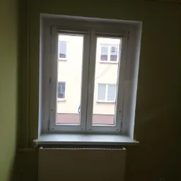 Białe, dwuskrzydłowe okno PCV z widokiem na blok mieszkalny, zamontowane w pokoju o ścianach pomalowanych na oliwkowy kolor, z grzejnikiem pod parapetem.