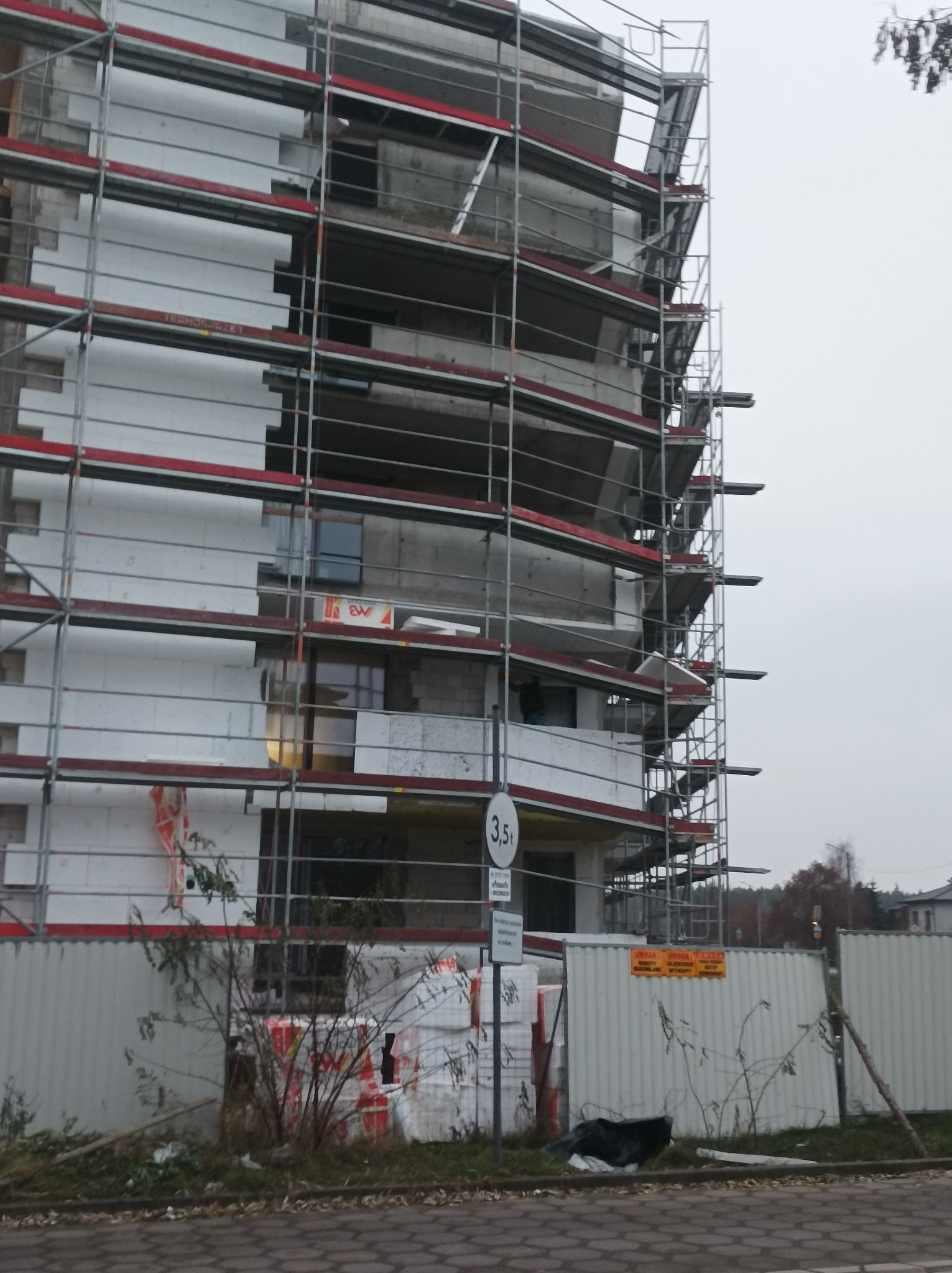 Wielopiętrowy budynek w trakcie prac elewacyjnych, z widocznym rusztowaniem i częściowo nałożonymi płytami izolacyjnymi, pod znakiem ograniczenia tonażu.