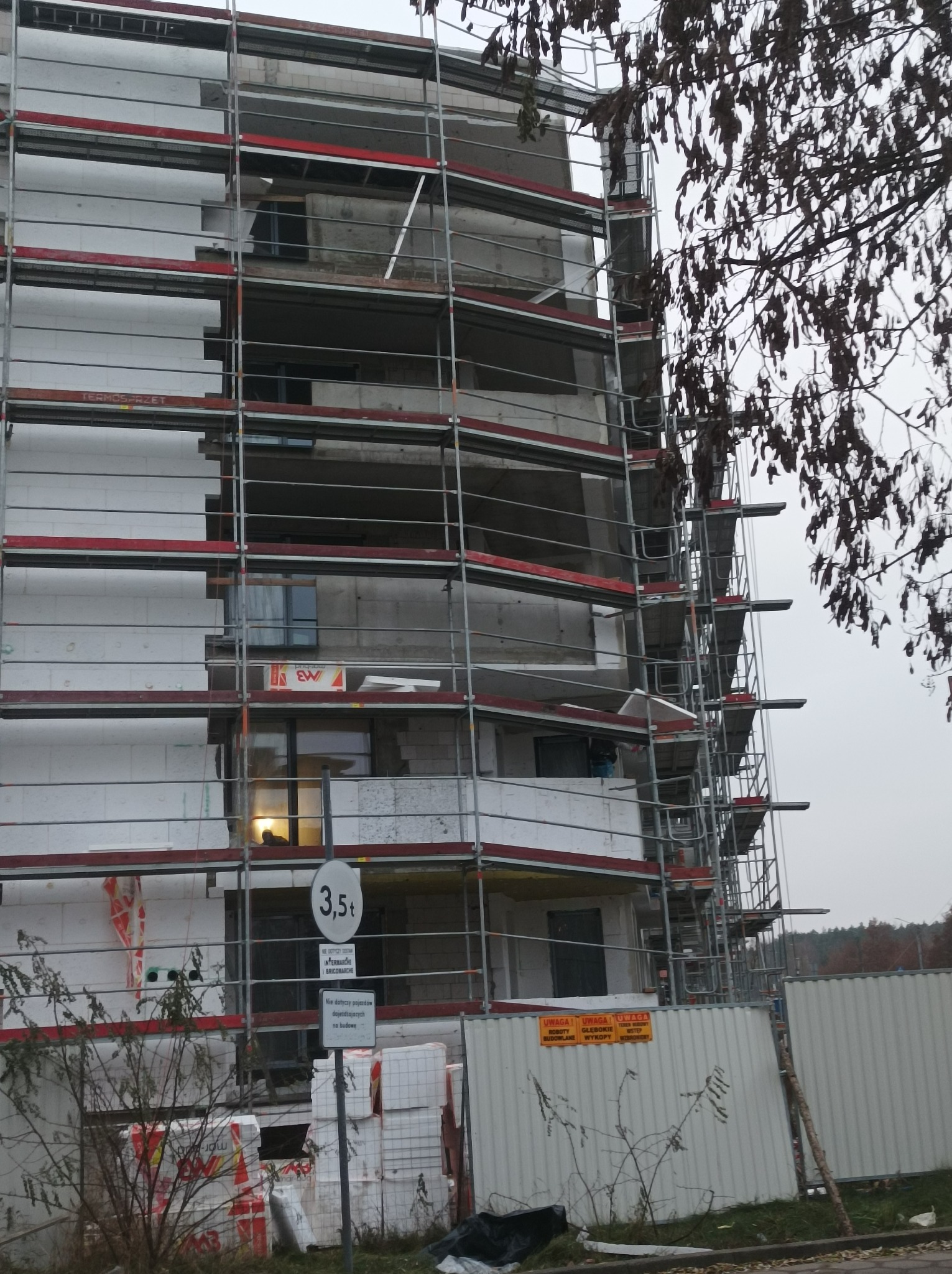 Budynek wielorodzinny w trakcie prac związanych z izolacją termiczną, widoczne rusztowania, płyty styropianowe, znak ograniczenia tonażu 3,5t oraz tablice ostrzegawcze.