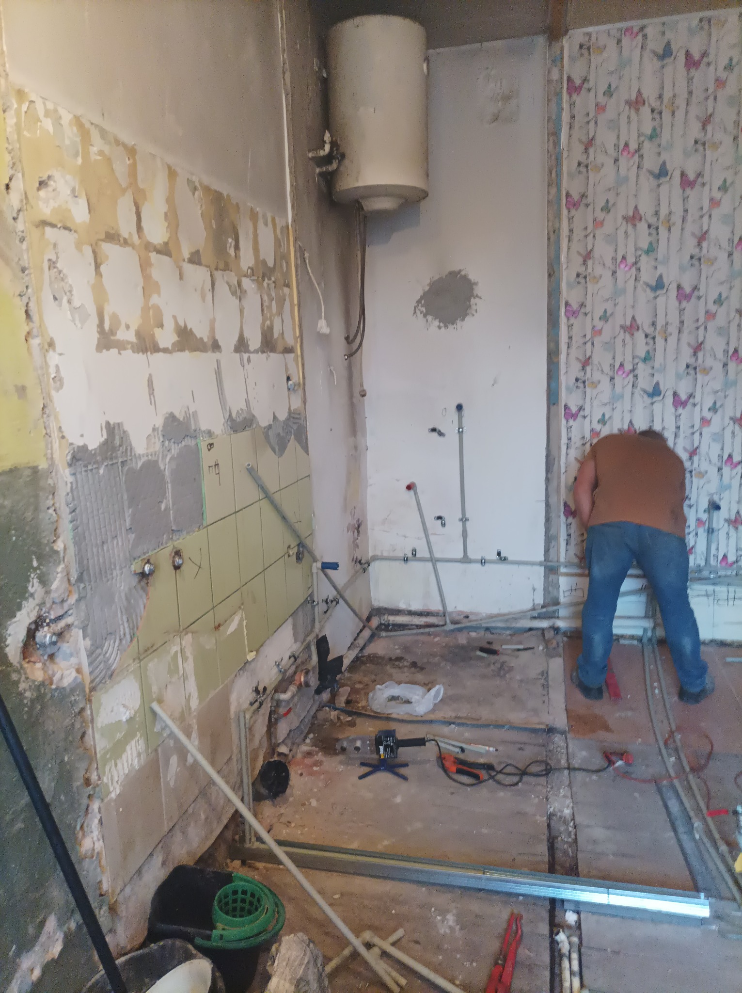 Demontaż starej glazury w kuchni podczas remontu, widoczne fragmenty płytek na ścianie, odsłonięte rury, narzędzia na podłodze i osoba w trakcie pracy.