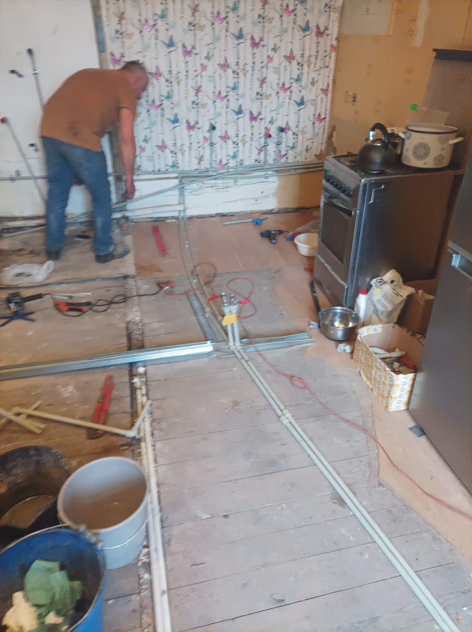 Pracownik instaluje rury grzewcze w trakcie remontu kuchni, widoczne narzędzia, stare deski podłogowe i ściana z tapetą w motyle.