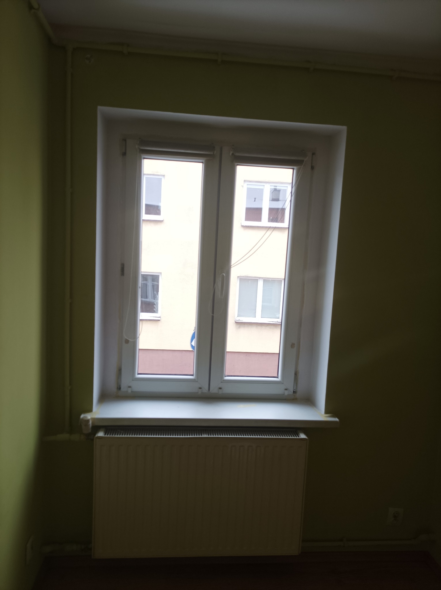 Białe, dwuskrzydłowe okno PCV z widokiem na blok mieszkalny, zamontowane w pokoju o ścianach pomalowanych na oliwkowy kolor, z grzejnikiem pod parapetem.