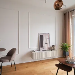 Jasne wnętrze pokoju dziennego z drewnianą podłogą, okrągłym stolikiem z kwiatami, dekoracyjnymi listwami na ścianie, wiszącą szafką z obrazem i rośliną doniczkową przy oknie.