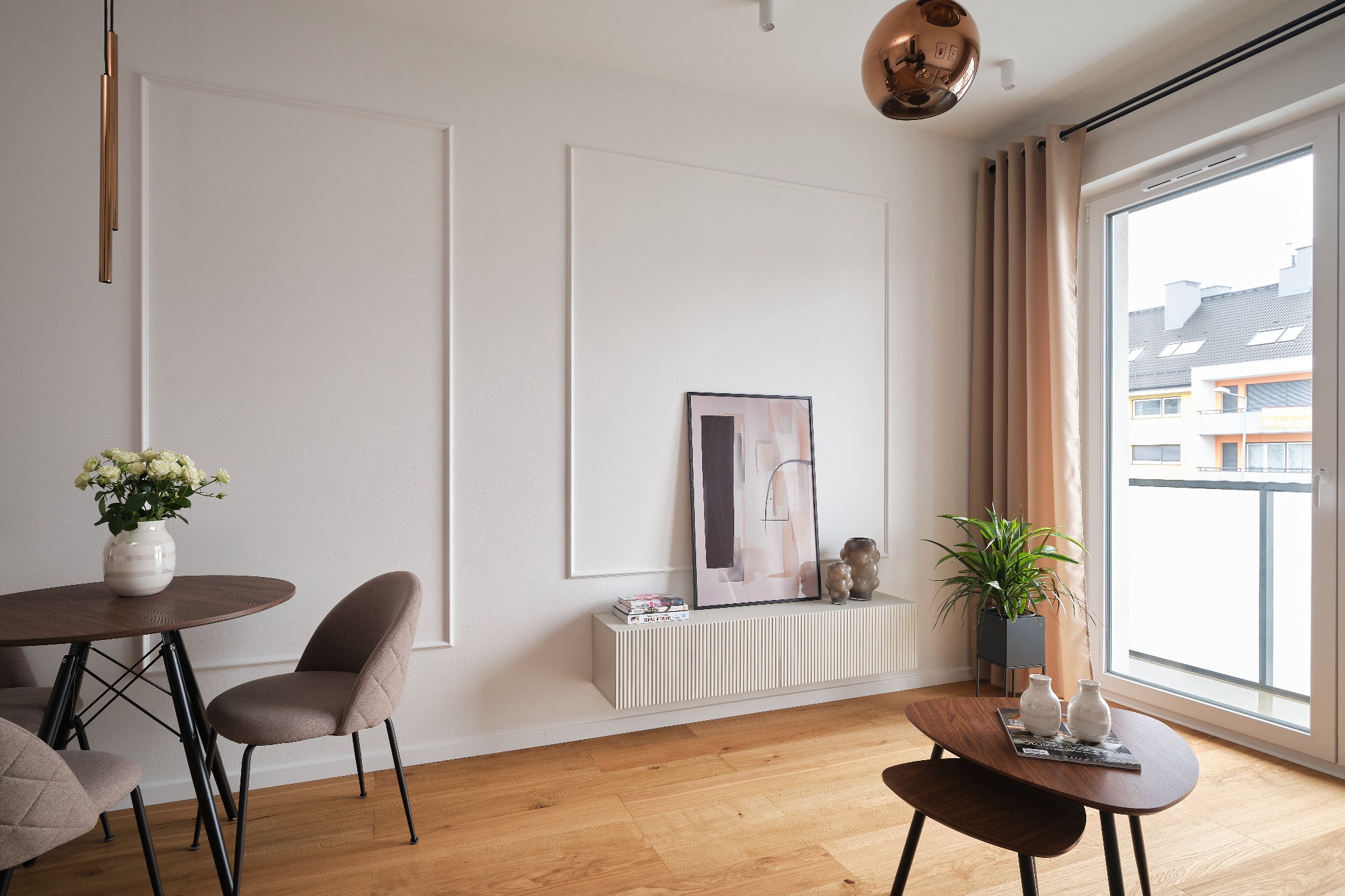 Jasne wnętrze pokoju dziennego z drewnianą podłogą, okrągłym stolikiem z kwiatami, dekoracyjnymi listwami na ścianie, wiszącą szafką z obrazem i rośliną doniczkową przy oknie.