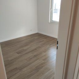 Klar Dom Andrzej Leszczyński - Pusty pokój z ułożonymi panelami podłogowymi w odcieniach brązu, widok zza uchylonych drzwi, białe ściany, okno z widokiem na zewnątrz, wisząca żarówka bez klosza.